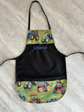 A black apron with a personalized name 'Charlie' in blue on the chest, featuring green fabric pockets at the bottom printed with colorful tractor illustrations.